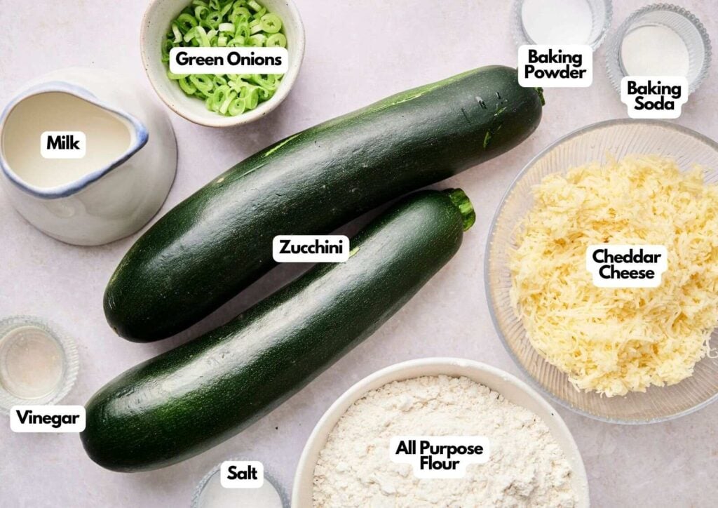 Various labeled ingredients for zucchini bread are arranged on a surface, including fresh zucchini, green onions, cheddar cheese, flour, milk, vinegar, salt, baking soda, and baking powder.