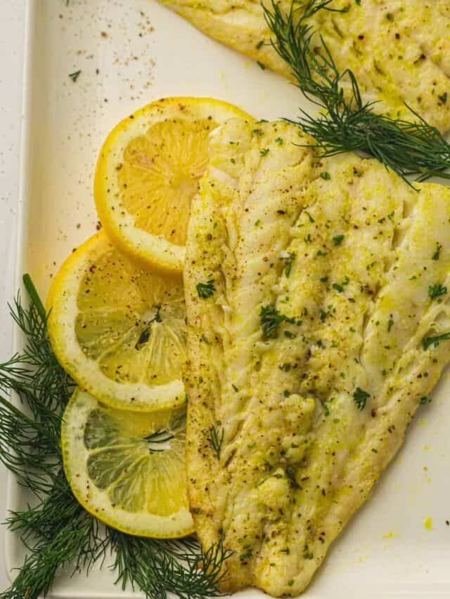 A seasoned, baked fish fillet garnished with lemon slices and fresh dill on a white plate.
