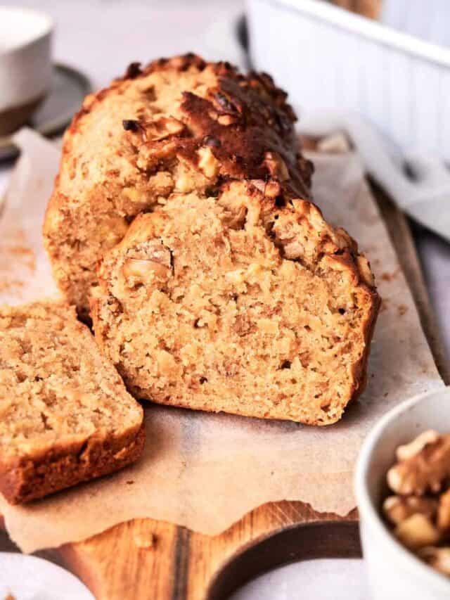 A loaf of banana bread topped with chopped nuts, partially sliced on a wooden cutting board with a bowl of nuts in the foreground and a cup in the background.