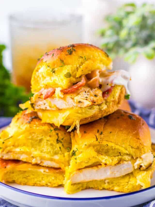 Three chicken sliders with cheese and bacon in golden buns are stacked on a plate, with a drink and greenery blurred in the background.