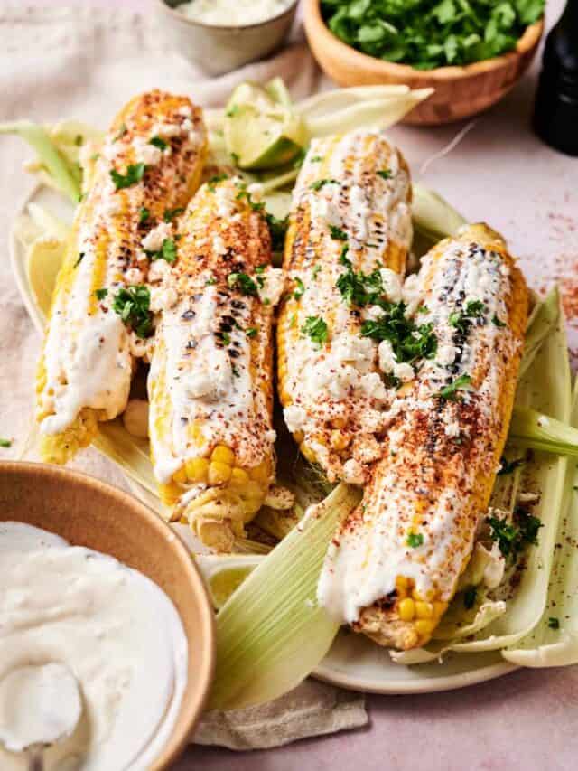 Four ears of grilled corn covered in mayonnaise, crumbled cheese, chili powder, and chopped cilantro are arranged on a plate. A bowl of white sauce, chopped cilantro, and crumbled cheese are nearby.