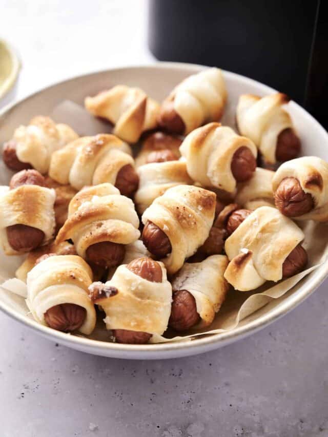 A white bowl filled with pigs in a blanket, showing mini sausages wrapped in golden-brown puff pastry on a white surface.