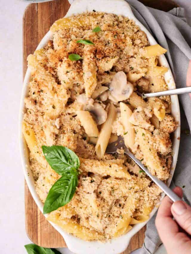 A baked pasta dish with breadcrumb topping, garnished with fresh basil, being served with a fork and spoon.