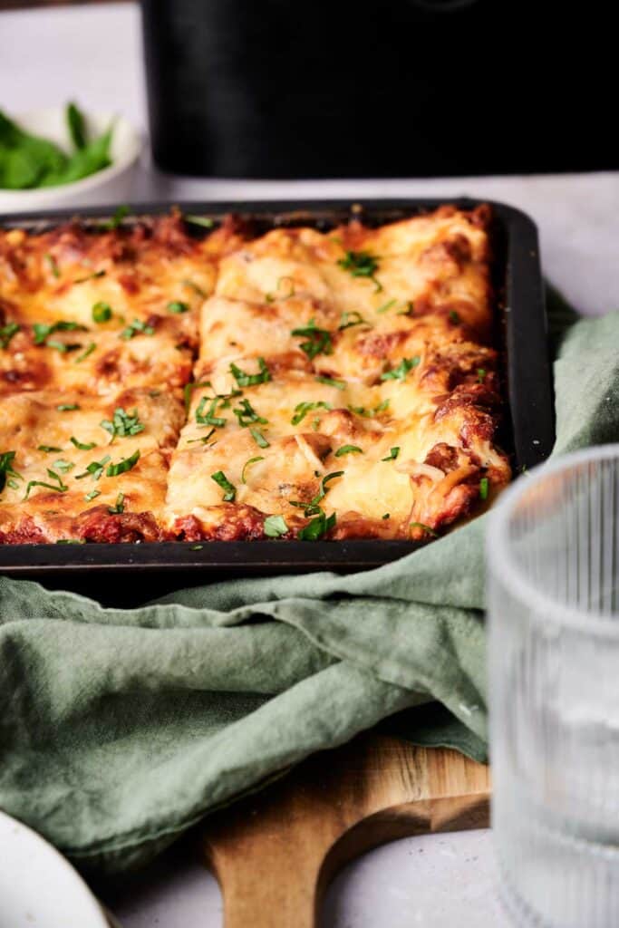 A tray of baked lasagna topped with melted cheese and garnished with chopped herbs on a green cloth.