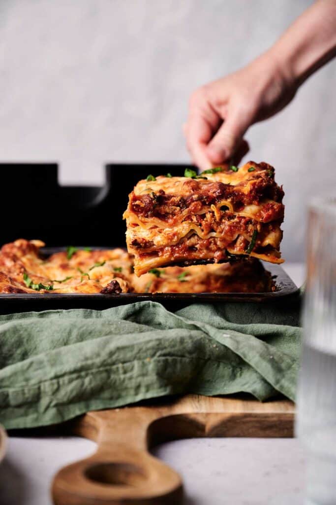 A person lifting a portion of lasagna from a baking dish, revealing layers of pasta, meat, and cheese.