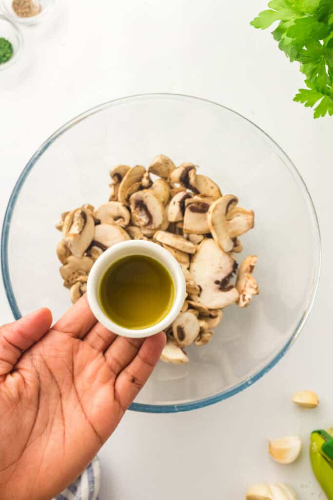 A hand holds a small cup of olive oil over a bowl of sliced mushrooms, perfect for making air fryer mushrooms. Parsley, garlic cloves, and seasonings are visible nearby on the countertop.