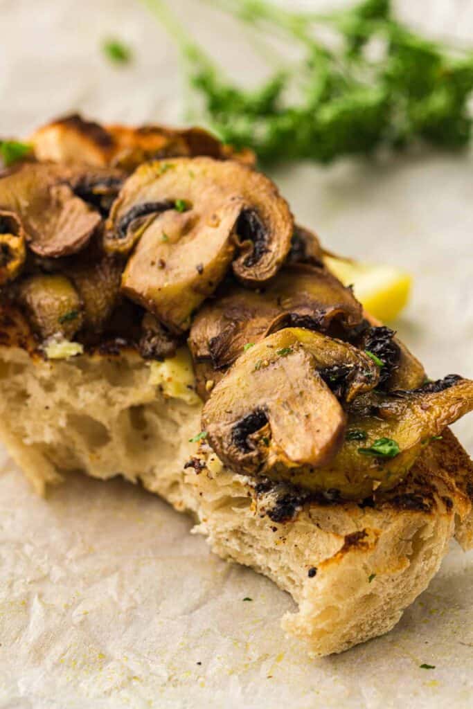 A slice of bread topped with air fryer mushrooms and herbs, partly eaten, placed on a sheet of parchment paper with a sprig of parsley in the background.