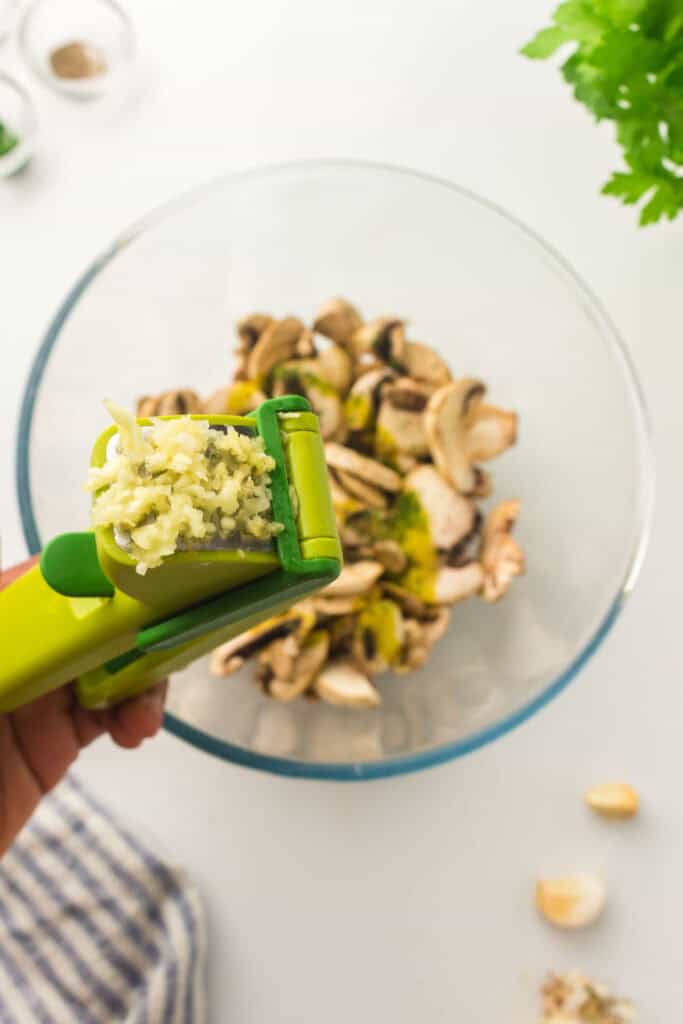 A garlic press releases minced garlic into a glass bowl containing sliced mushrooms, with additional garlic cloves and fresh greenery nearby—perfect for preparing air fryer mushrooms.