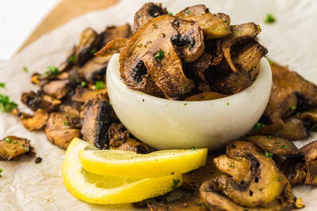 A small white bowl filled with air fryer mushrooms, garnished with chopped herbs, with lemon slices placed in front.