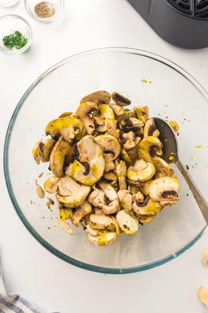 A glass bowl filled with air fryer mushrooms mixed with seasonings. A spoon is placed in the bowl, and small bowls of additional seasonings are visible nearby.
