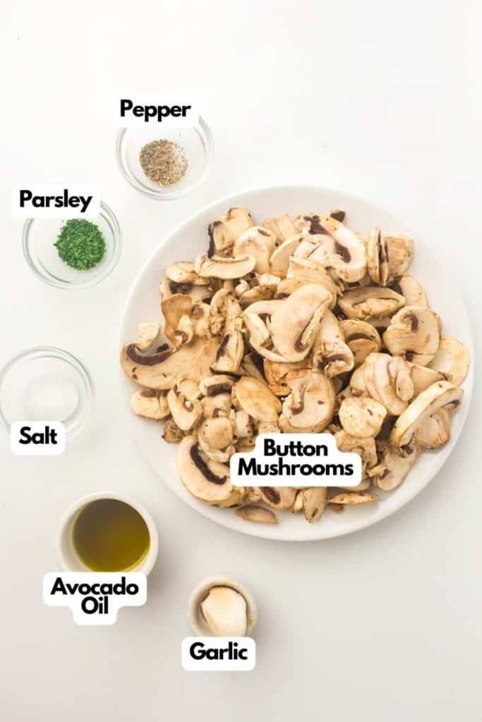 A plate of sliced button mushrooms is surrounded by small bowls of parsley, salt, pepper, avocado oil, and a clove of garlic—all labeled and ready to make delicious air fryer mushrooms.