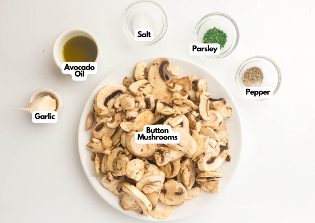 A white plate of sliced button mushrooms, perfect for making air fryer mushrooms, is surrounded by small bowls of avocado oil, garlic, salt, parsley, and pepper on a white surface.