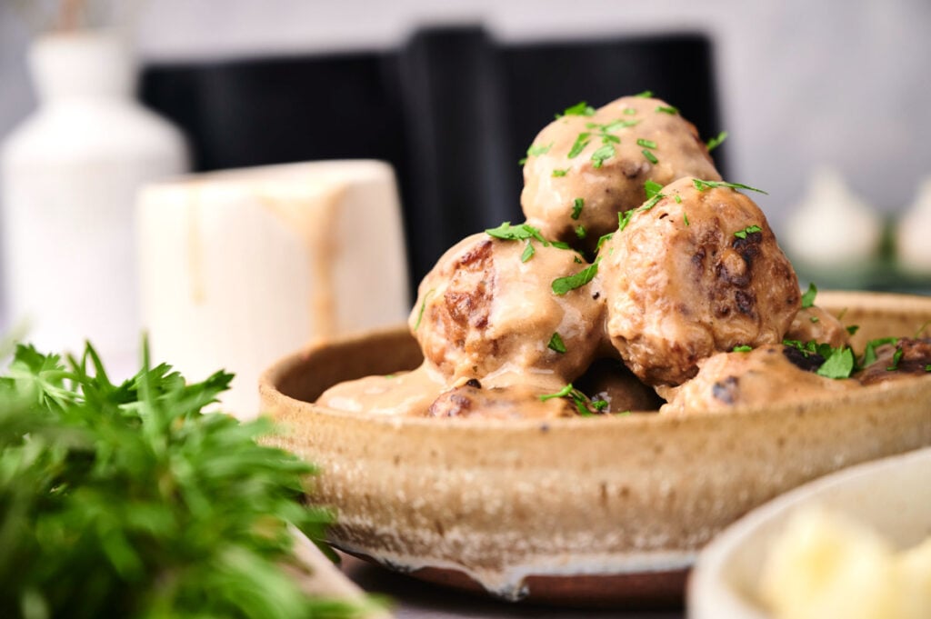 A close-up of a ceramic bowl filled with air fryer Swedish meatballs covered in creamy sauce, garnished with chopped parsley.