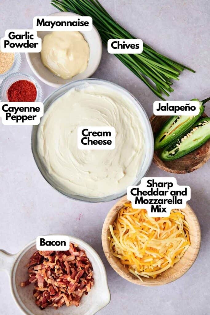 Ingredients for Bacon Jalapeno Popper Cheese Balls arranged on a surface: cream cheese, sharp cheddar and mozzarella mix, bacon, garlic powder, cayenne pepper, mayonnaise, chives, and a halved jalape&ntilde;o.