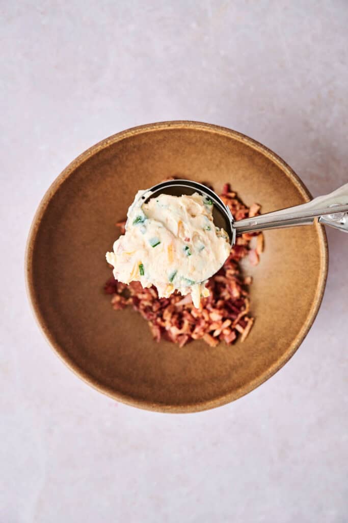 A scoop of creamy mixture is being held above a bowl containing chopped ingredients, ready to transform into delicious bacon jalapeno popper cheese balls.