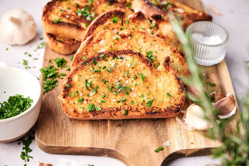 Four slices of garlic bread garnished with chopped parsley and coarse salt rest on a wooden cutting board, surrounded by bowls of fresh herbs and salt.