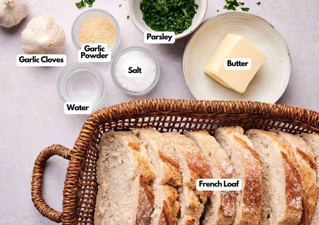 Wicker basket with sliced French bread loaf, butter, chopped parsley, garlic cloves, garlic powder, salt, and water on a countertop—perfect ingredients for making delicious homemade garlic bread.