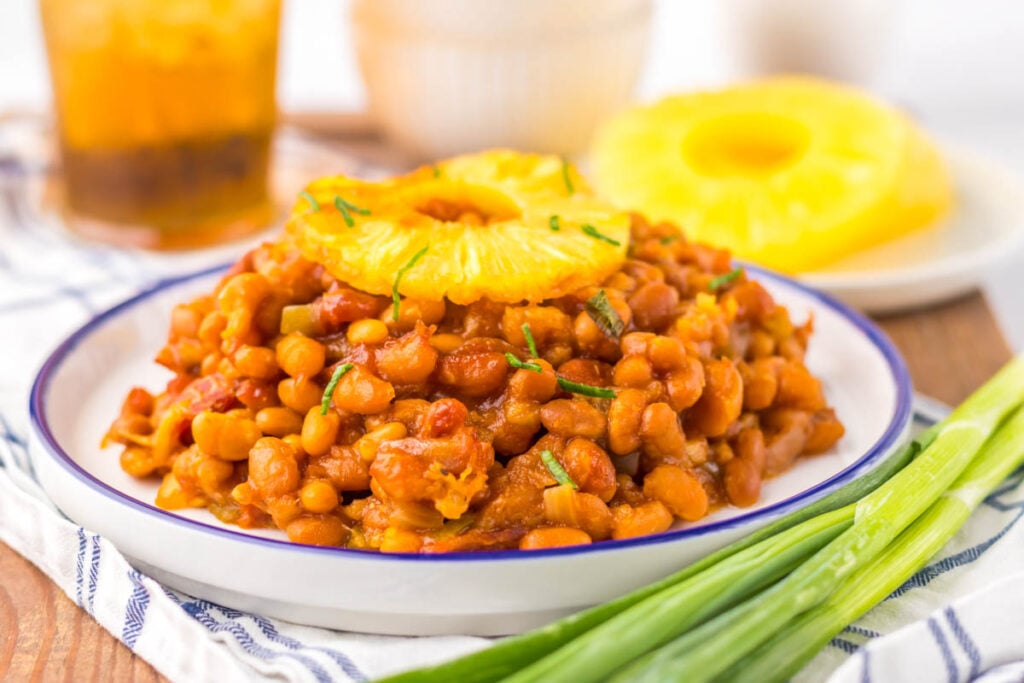 A plate of Hawaiian baked beans topped with pineapple rings, with green onions beside the plate and a drink and sliced pineapple in the background.