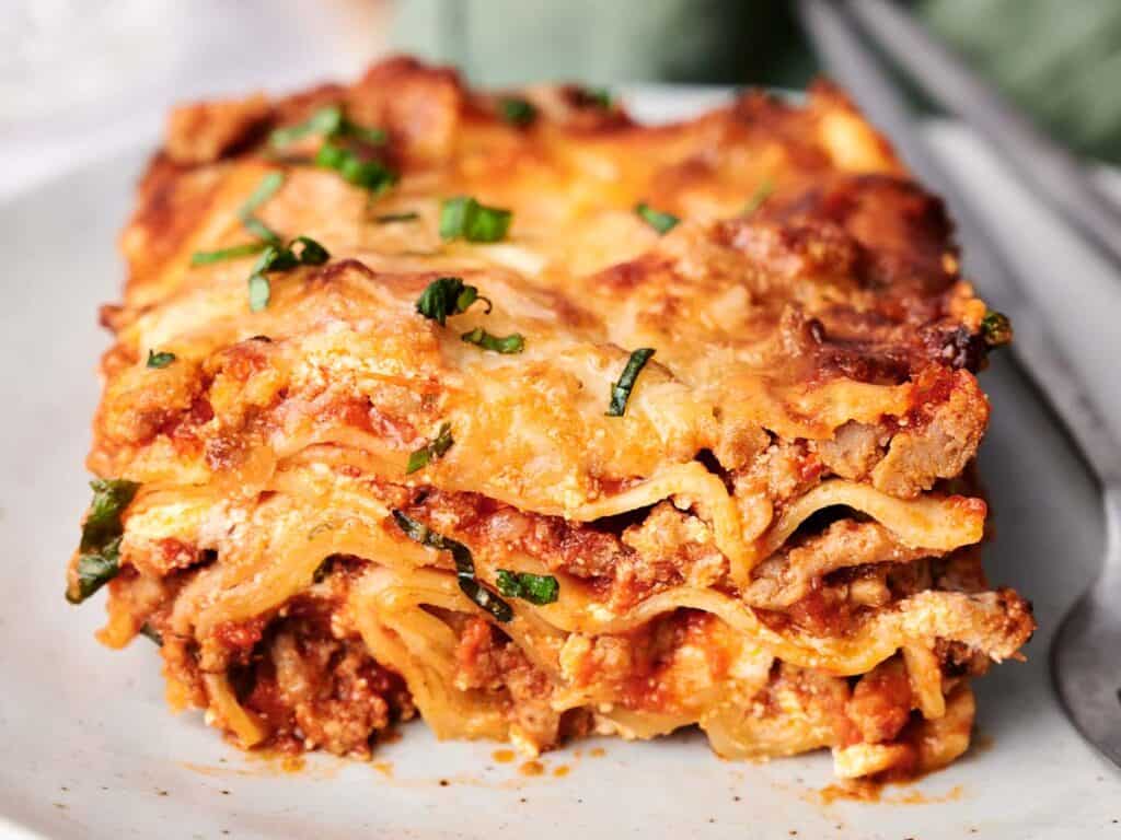 A close-up of a slice of lasagna with visible layers of pasta, meat sauce, and cheese, garnished with chopped herbs.