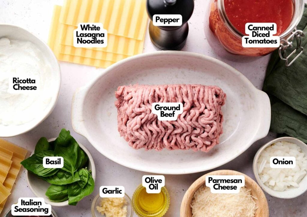 Top-down view of air fryer lasagna ingredients including ground beef, white lasagna noodles, canned diced tomatoes, onion, ricotta cheese, parmesan cheese, basil, garlic, olive oil, Italian seasoning, and pepper.