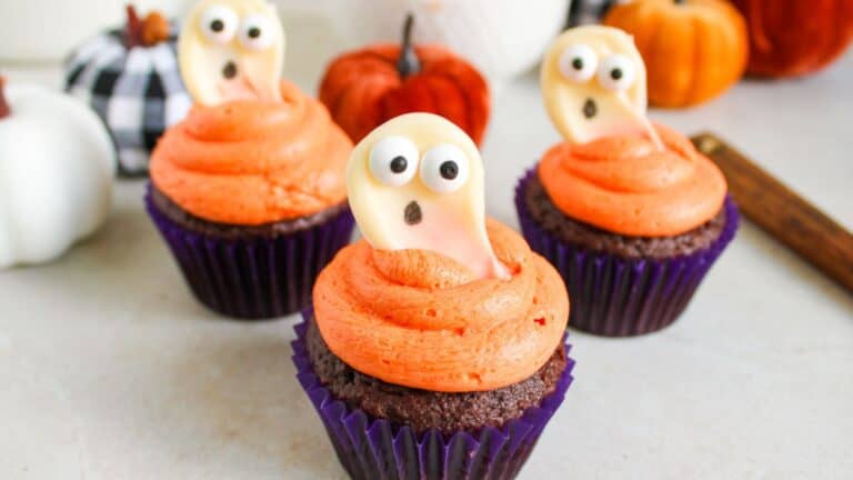 Three chocolate cupcakes with orange frosting are topped with white ghost-shaped decorations featuring googly eyes and open mouths. Pumpkins and fall decor are in the background.