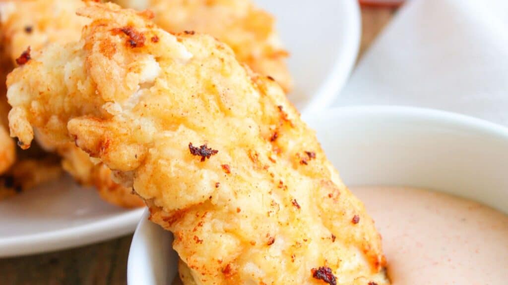 Close-up image of a crispy fried chicken tender being dipped into a creamy sauce, with more chicken tenders in the background.