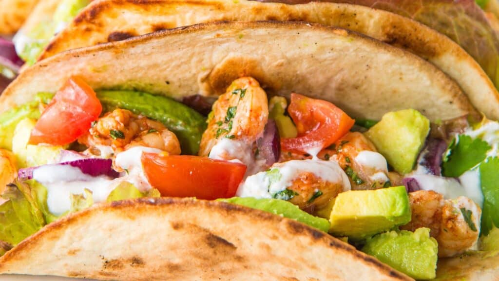 Close-up of a taco filled with grilled shrimp, diced tomatoes, avocado chunks, red onion, and drizzled with a white sauce. The taco shell appears toasted and crispy.
