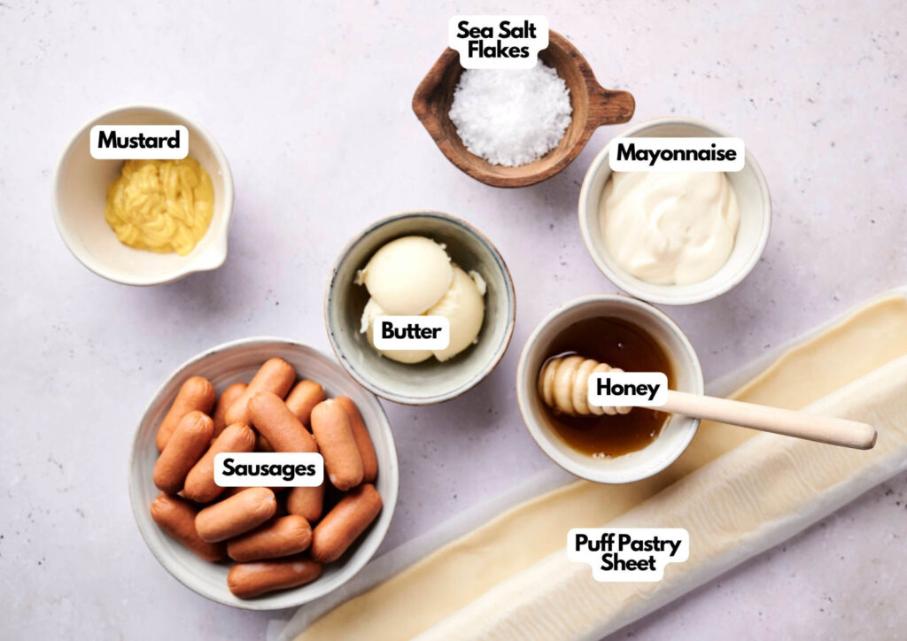 Top-down view of bowls containing mustard, butter, sea salt flakes, mayonnaise, honey, sausages, and a rolled-out puff pastry sheet—perfect ingredients for making air fryer pigs in a blanket on a light surface.