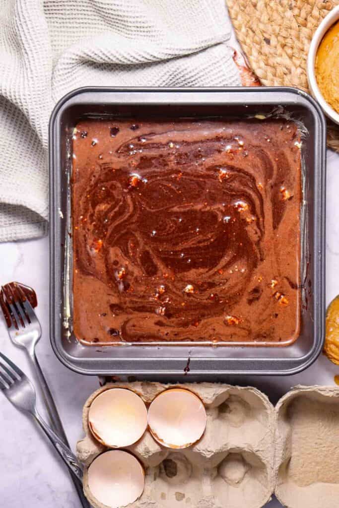 A baking pan filled with chocolate swirl batter surrounded by baking utensils, a dishcloth, a bowl, and broken eggshells on a countertop.