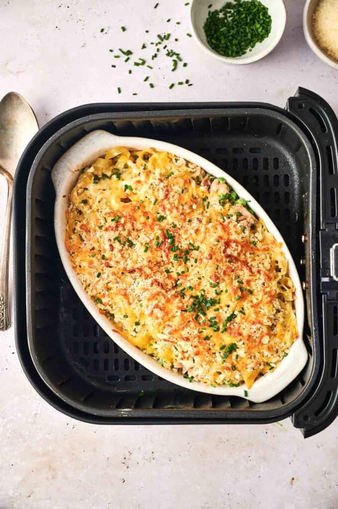 A casserole with a crispy top is cooking inside an air fryer basket. It is sprinkled with chopped chives. A bowl of extra chives is in the corner, and utensils are placed beside the air fryer.
