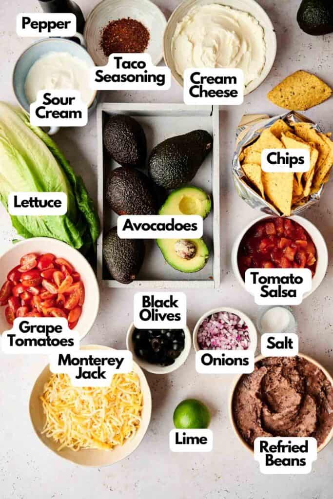 Ingredients for a 7 Layer Dip, including pepper, taco seasoning, cream cheese, sour cream, lettuce, avocados, chips, tomato salsa, onions, salt, refried beans, lime, Monterey Jack cheese, black olives and grape tomatoes.