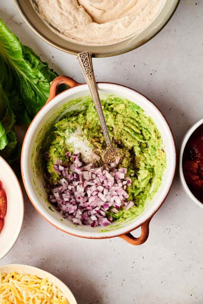 A bowl of guacamole with chopped red onions, salt, and pepper on top sits invitingly, reminiscent of a 7 Layer Dip. A spoon is placed in the bowl, ready for scooping. Surrounding the bowl are various fresh food ingredients.