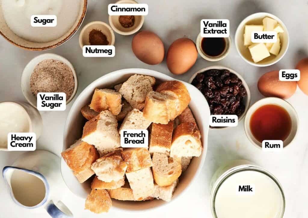 Ingredients for bread pudding arranged on a countertop, including French bread, eggs, milk, heavy cream, sugar, vanilla sugar, cinnamon, nutmeg, vanilla extract, butter, raisins, and rum&mdash;a classic setup for homemade bread pudding.