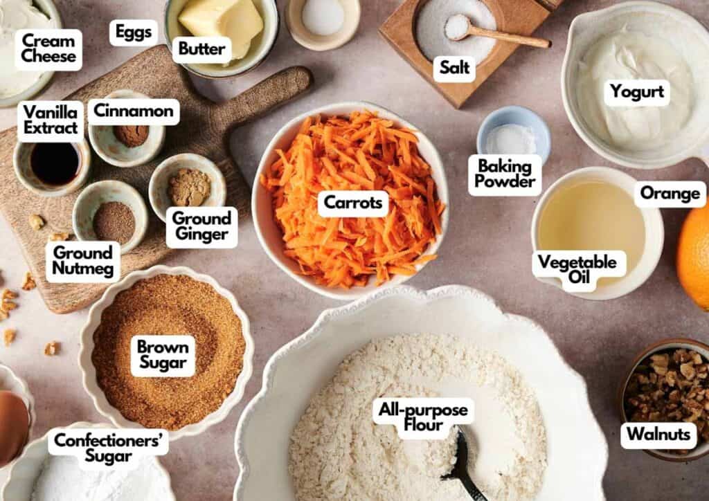 Assorted labeled carrot cake ingredients, including grated carrots, flour, sugars, yogurt, eggs, butter, spices, baking powder, oil, walnuts, cream cheese, orange, and vanilla extract.