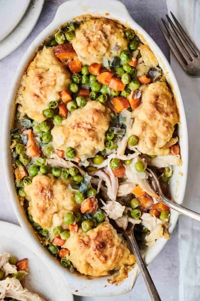 A white baking dish with chicken and vegetables topped with golden-brown biscuits, reminiscent of a delicious chicken and dumpling casserole, served with two metal serving spoons.