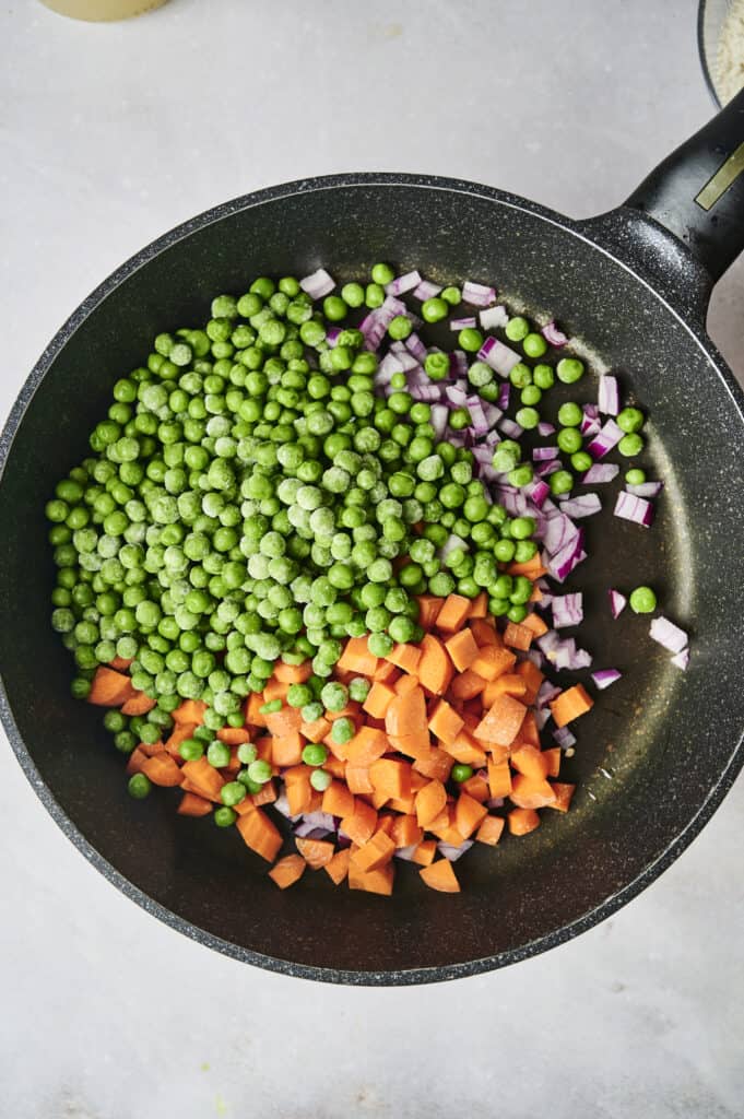A frying pan containing diced carrots, red onions, and green peas, ready to be cooked for a hearty chicken and dumpling casserole.