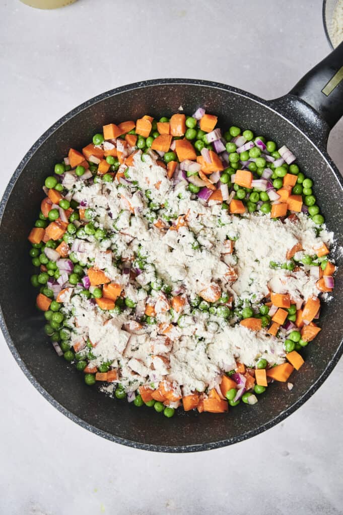 A black frying pan contains a delightful mixture of green peas, diced carrots, and chopped red onions with a sprinkling of flour on top, reminiscent of the hearty filling in a chicken and dumpling casserole.