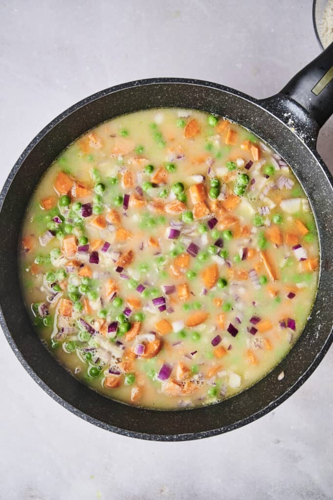 A pan filled with a colorful mixture of peas, diced carrots, red onions, and other vegetables in a creamy liquid forms the base of this delicious chicken and dumpling casserole.
