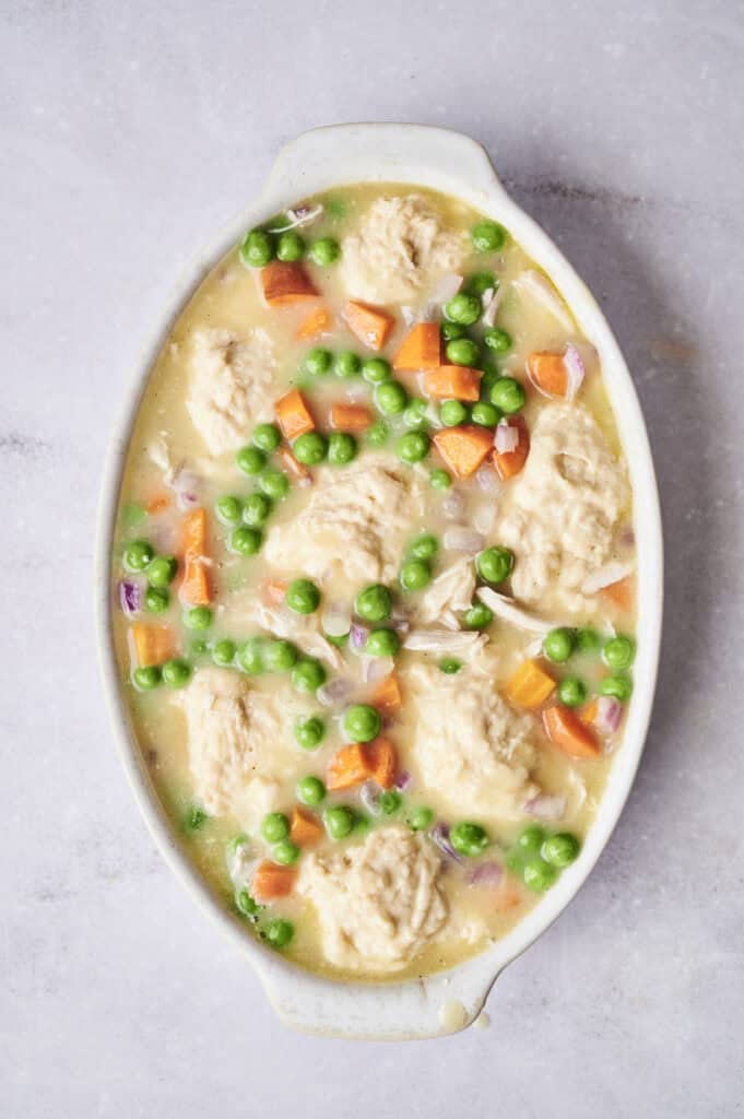 An oval baking dish filled with a hearty chicken and dumpling casserole, brimming with green peas, carrot chunks, and chopped onions in a creamy sauce.