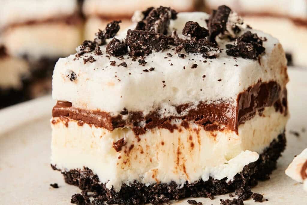 A close-up of a decadent Chocolate Lasagna dessert bar with a crumbly chocolate cookie base, creamy white and chocolate layers, and crushed cookies on top.