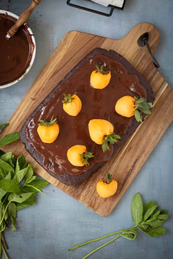 A wooden board with a rectangular Chocolate Pumpkin Patch Brownie Cake topped with glossy chocolate ganache and six strawberries coated in orange icing. Fresh mint leaves are placed on the board. A bowl of chocolate sauce is nearby.
