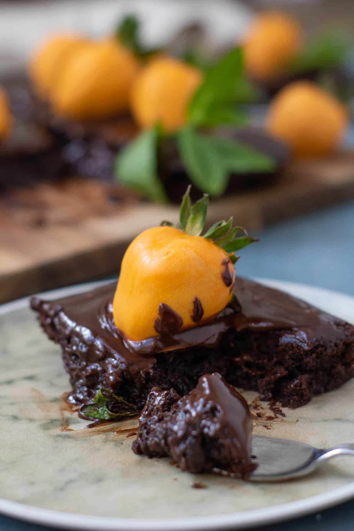 A slice of chocolate pumpkin patch brownie cake topped with a chocolate-covered yellow fruit, served on a plate with a fork beside it. More fruits and pieces of cake are blurred in the background.