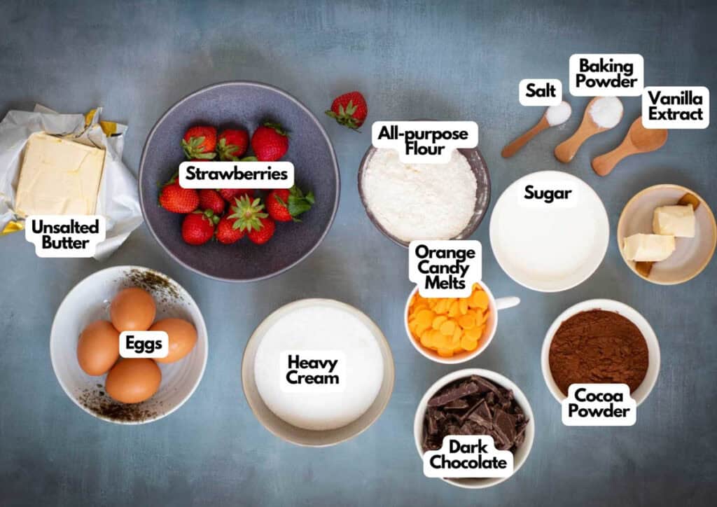 Various baking ingredients for a Chocolate Pumpkin Patch Brownie Cake, including strawberries, eggs, butter, cream, flour, orange candy melts, chocolate, cocoa powder, sugar, salt, baking powder, and vanilla extract arranged on a table.