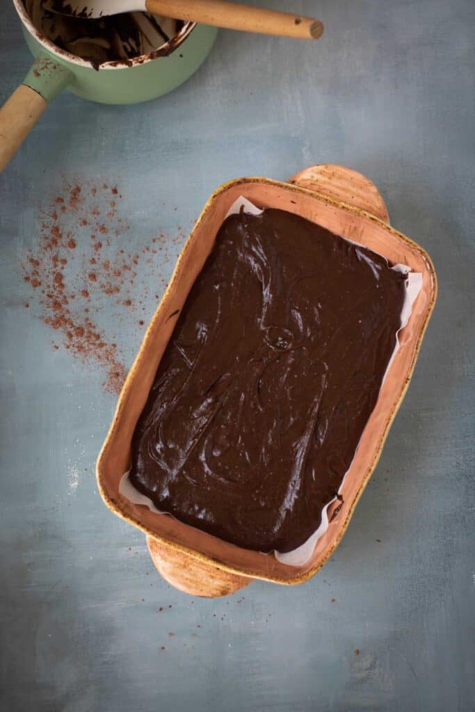 A rectangular baking dish filled with Chocolate Pumpkin Patch Brownie Cake batter is placed on a teal surface. A saucepan and a wooden spoon are in the upper left corner, with cocoa powder scattered nearby.