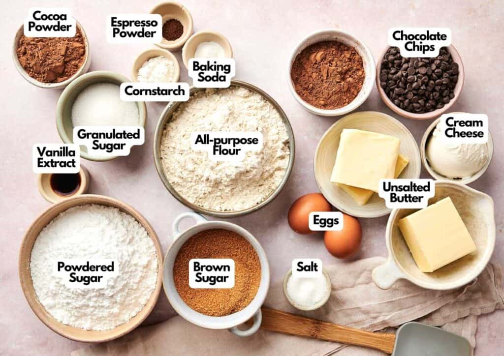 Various baking ingredients, perfect for making a Chocolate Pumpkin Patch Brownie Cake, including flour, sugars, cocoa powder, chocolate chips, baking soda, cornstarch, espresso powder, vanilla extract, butter, cream cheese, eggs, and salt are arranged on a counter.