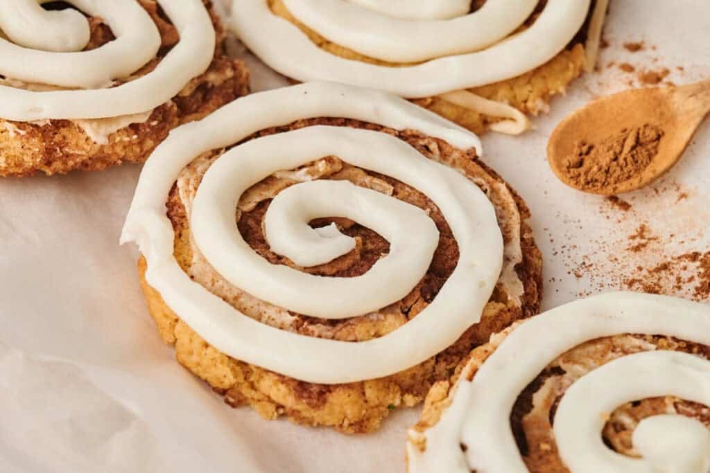 Close-up of cinnamon roll cookies topped with white icing swirls, placed on parchment paper next to a wooden spoon with ground cinnamon&mdash;perfect alongside a slice of Chocolate Pumpkin Patch Brownie Cake.