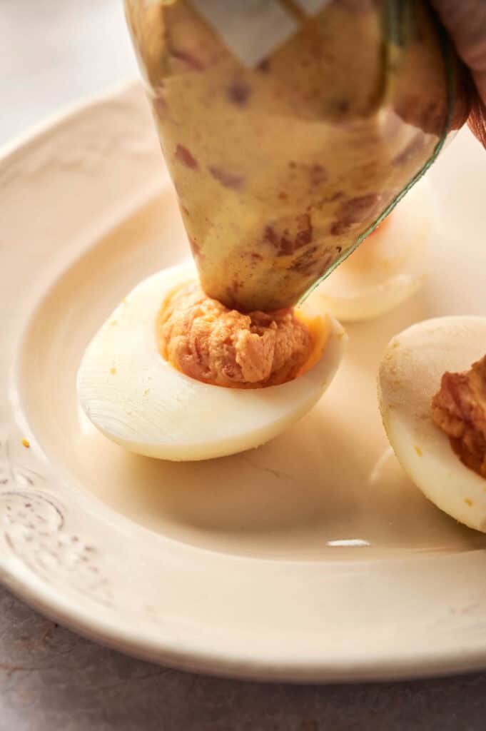 Close-up of a piping bag deftly filling deviled eggs with a creamy mixture on a beige plate.