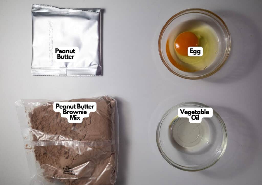 A packet of peanut butter, a bowl with an egg, a bag of Chocolate Pumpkin Patch Brownie Cake mix, and a bowl of vegetable oil are arranged on a white surface.