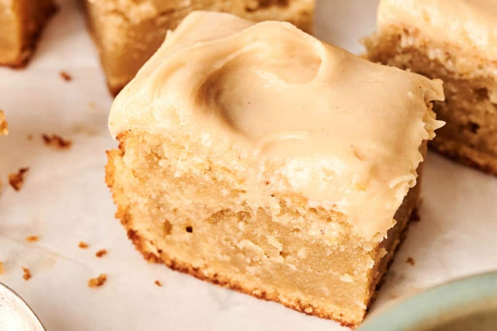 A close-up of a thick square piece of Chocolate Pumpkin Patch Brownie Cake, topped with creamy light brown frosting, sitting on a white surface.