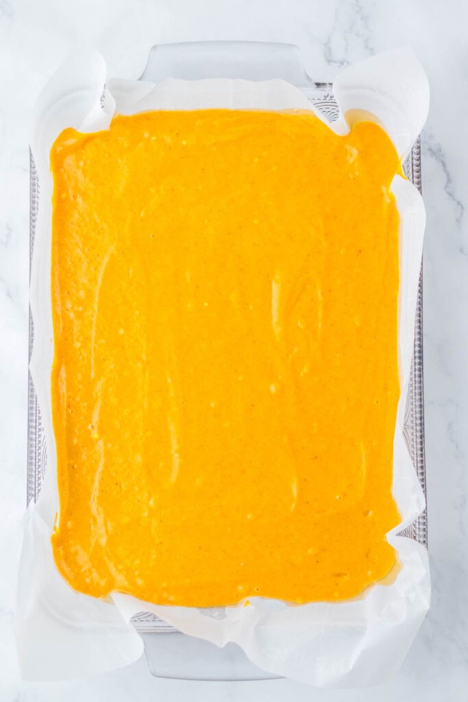 A rectangular baking dish lined with parchment paper is filled with smooth, orange-colored Pumpkin Caramel Cake batter, ready to be baked.